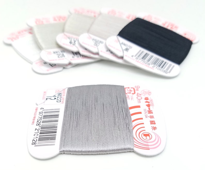 May include: A spool of silver thread with a white label that reads "ワイヤー絹手縫糸 9号 40m プラ 100% Sik" and a barcode. The label also has a red circle with a white letter "C" inside.