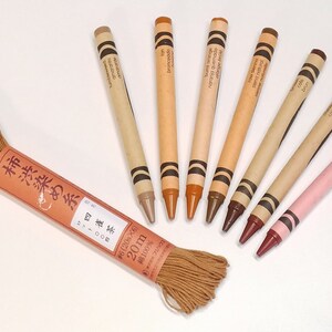 May include: A set of eight brown and pink crayons with Japanese text on the labels. The crayons are arranged in a semi-circle around a spool of brown string. The string is labeled in Japanese with the text "柿渋染め糸雀 371000 #6 (206&times;6) 20m 綿100%"