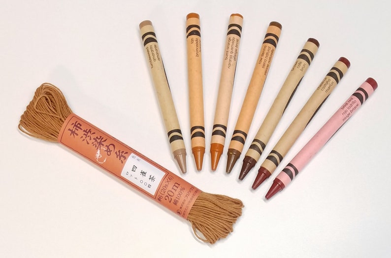 May include: A set of eight brown and pink crayons with Japanese text on the labels. The crayons are arranged in a semi-circle around a spool of brown string. The string is labeled in Japanese with the text "柿渋染め糸雀 371000 #6 (206&times;6) 20m 綿100%"
