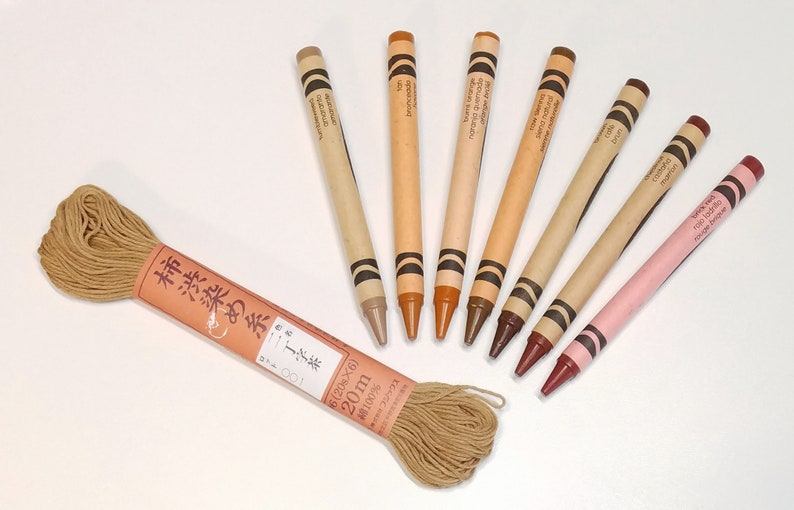 May include: A set of seven brown crayons arranged in a semi-circle with a spool of brown thread and a label that reads "柿渋染め糸子茶 100 6(20s&times;6) 20m 100% send tunal cotto&ntilde;o amarato Bronceado".