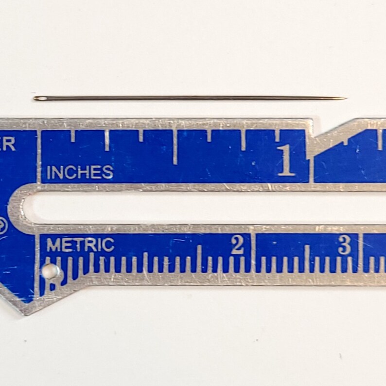 May include: A close-up of a silver sewing needle measuring 1 inch in length against a blue ruler with inch and metric markings.