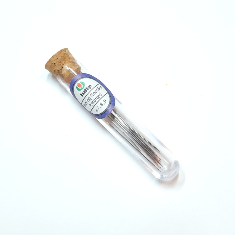 May include: A clear glass tube with a cork top containing a variety of sewing needles. The label on the tube reads "Tulip Sewing Needles Assorted #7, 8, 9".
