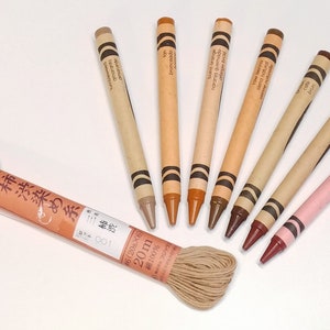 May include: A set of eight crayons in various shades of brown, beige, and pink. The crayons are arranged in a semi-circle with the tips pointing towards the center. The crayons are labeled with their colors in Japanese and English. A spool of brown thread is in the center of the semi-circle.