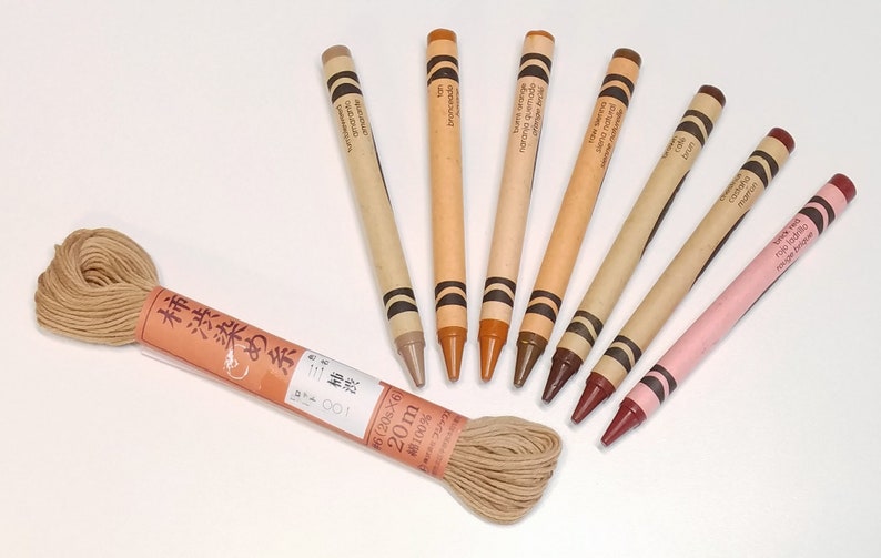 May include: A set of eight crayons in various shades of brown, beige, and pink. The crayons are arranged in a semi-circle with the tips pointing towards the center. The crayons are labeled with their colors in Japanese and English. A spool of brown thread is in the center of the semi-circle.