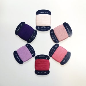 May include: Six spools of thread in various shades of pink, purple, and red. Each spool is labelled "SASHIKO" and has details about the thread, including the material, size, and length.