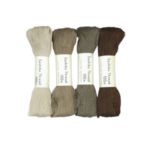 Könnte beinhalten: Vier Spulen Sashiko-Garn, jede 100 m lang. Die Spulen sind in Beige-, Braun- und Grautönen. Das Etikett auf jeder Spule lautet "Sashiko Thread 100m Cotton 100% Made in Japan".