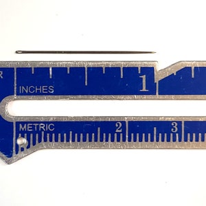 May include: A close-up of a silver ruler with blue markings. The ruler has both inches and metric measurements. A small, thin, silver needle is placed on top of the ruler, measuring approximately 1 inch in length.
