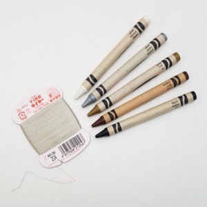 May include: Six wooden crayons with paper wrappers. The crayons are white, silver, gold, brown, and black. The wrappers have the color names in both English and Spanish. The crayons are arranged in a row on a white surface.