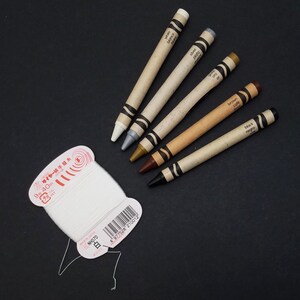 May include: Five crayons in a variety of colors, including white, silver, gold, brown, and black. The crayons are labeled with their color in both English and Spanish. The crayons are on a black surface.
