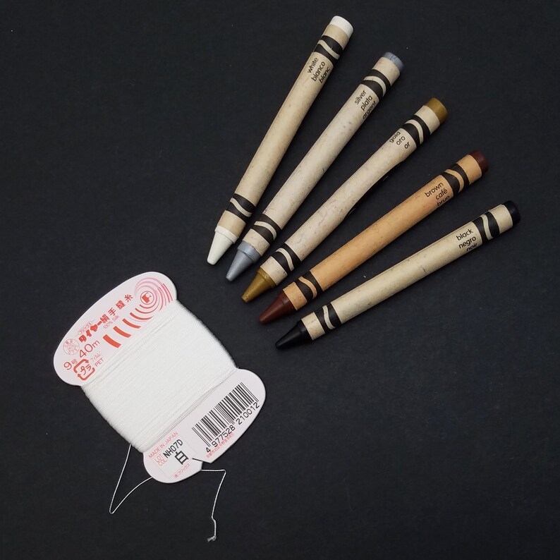 May include: Five crayons in a variety of colors, including white, silver, gold, brown, and black. The crayons are labeled with their color in both English and Spanish. The crayons are on a black surface.