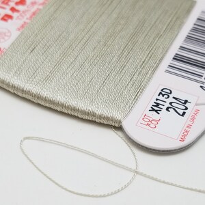 May include: A spool of light gray thread with a white label that reads "LOT XM13D COL 204 MADE IN JAPAN".