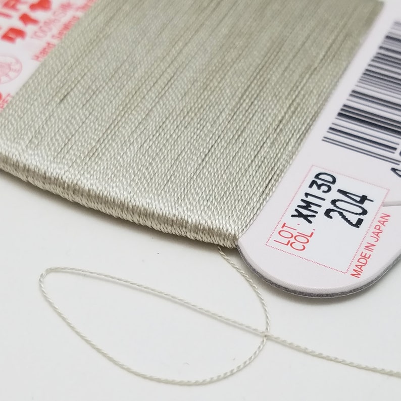 May include: A spool of light gray thread with a white label that reads "LOT XM13D COL 204 MADE IN JAPAN".
