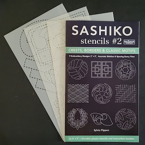 May include: A set of three plastic Sashiko stencils for embroidery, featuring 9 designs, including crests, borders, and classic motifs. The stencils are 5 inches by 9 inches and come with an instruction booklet. The booklet has a black cover with white text that reads "Sashiko stencils #2 Fast2Mark Tools Crests, Borders & Classic Motifs 9 Embroidery Designs 3" x 5", Accurate Stitches & Spacing Every Time Sylvia Pippen (3) 5" x 9" - Durable plastic stencils and instruction booklet".