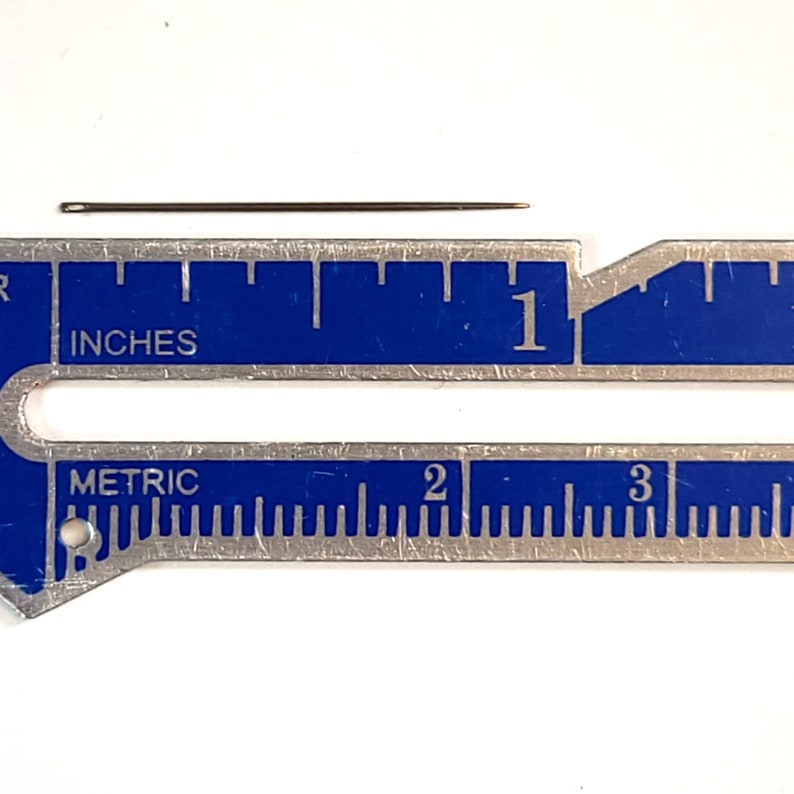 May include: A close-up of a silver ruler with blue markings. The ruler has both inches and metric measurements. A small, thin needle is placed on top of the ruler, measuring approximately 1 inch in length.