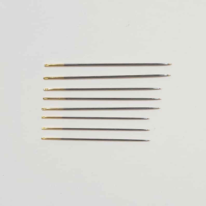 May include: A set of eight silver sewing needles with gold tips. The needles are arranged vertically in a row.