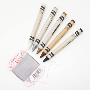 May include: A set of six crayons in a variety of colors, including white, silver, gold, brown, and black. The crayons are labeled with their color names in English, Spanish, and French. The crayons are arranged in a row on a white surface. A spool of gray thread is also visible in the image.