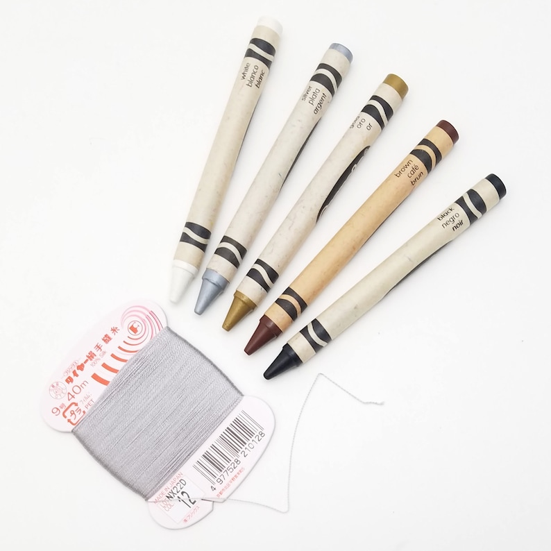 May include: A set of six crayons in a variety of colors, including white, silver, gold, brown, and black. The crayons are labeled with their color names in English, Spanish, and French. The crayons are arranged in a row on a white surface. A spool of gray thread is also visible in the image.