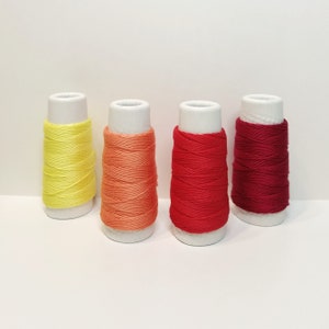 May include: Four spools of thread in yellow, orange, red, and burgundy colors. The spools are wound with thread and are sitting on a white surface.