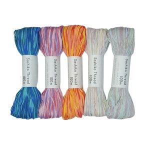 Könnte beinhalten: Fünf Spulen mit farbigem Sashiko-Garn, jede 100 Meter lang. Die Farben sind blau und grün, rosa und weiß, orange und gelb, hellblau und weiß sowie lila und weiß.