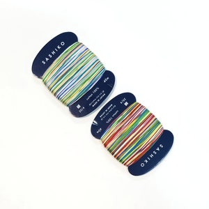 May include: Two spools of Sashiko thread with colorful stripes. Each spool is labeled with "SASHIKO", "cotton 100%", and "MADE IN JAPAN". The thread is 40m long. The spools are on a white background.