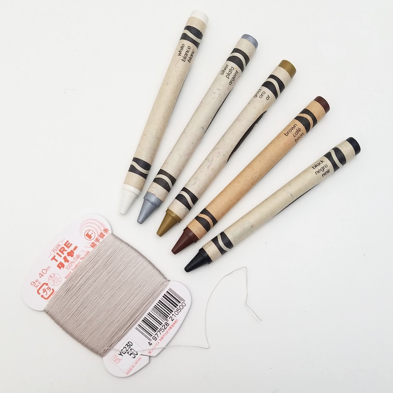 May include: A spool of gray thread with a label that reads "TIRE 40m 100% Silk" and a set of six crayons in various colors, including white, silver, gold, brown, and black.