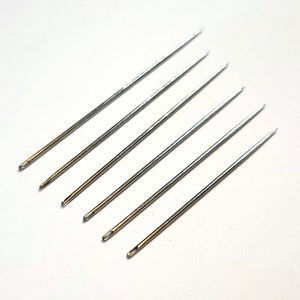 May include: Six silver sewing needles with gold eye holes. The needles are arranged in a fan shape on a white background.