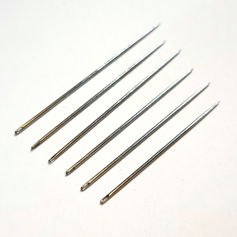 May include: Six silver sewing needles with gold eye holes. The needles are arranged in a fan shape on a white background.