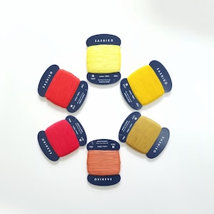 Puede incluir: Seis carretes de hilo de bordar en varios colores, incluyendo rojo, naranja, amarillo y marrón. Cada carrete está etiquetado con "SASHIKO" e incluye información sobre el contenido de algodón y el tamaño.