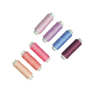 May include: A selection of colourful thread spools in shades of pink, purple, and blue. The spools are arranged diagonally against a white background, showcasing the variety of colours available for sewing and crafting projects.