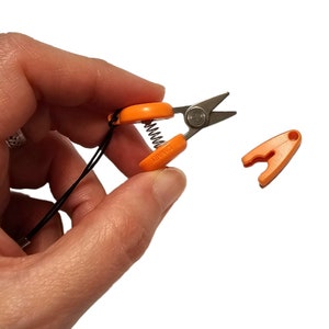 Puede incluir: Pequeñas tijeras naranjas y plateadas con un resorte y un cordón negro. Las tijeras tienen mangos naranjas con la palabra "ZANARY" impresa. También se ve una pieza de plástico naranja separada.