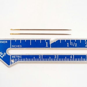 May include: Two gold-eyed needles measuring 3.8 cm long, placed on a blue and silver ruler with inch and metric markings.