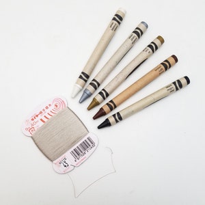 May include: Five wooden crayons in different colors: white, silver, gold, brown, and black. The crayons are labeled in English, Spanish, and French. A spool of beige thread is also in the image.