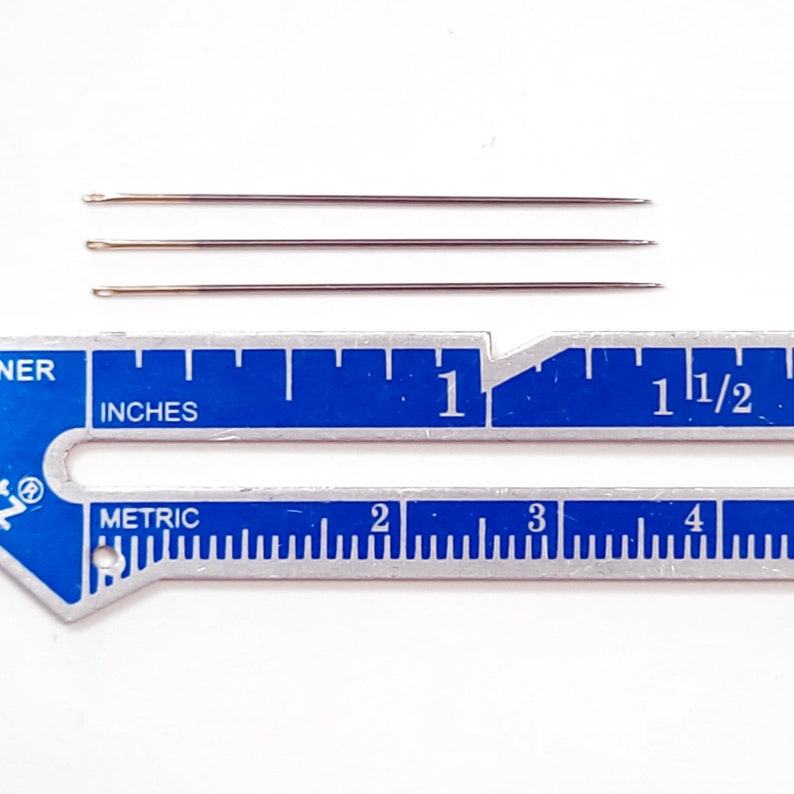 May include: Three silver sewing needles measuring 1 inch in length, displayed against a blue and silver ruler.