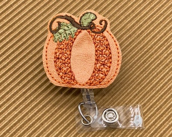 May include: Orange pumpkin-shaped fabric badge reel with a green stem and brown leaves. The reel is attached to a clear plastic retractable badge holder.