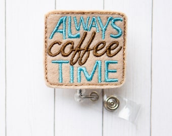 May include: A beige felt badge holder with the text "ALWAYS coffee TIME" embroidered in blue and brown thread. The badge holder is attached to a clear plastic retractable badge reel with a silver swivel clip.