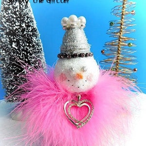 May include: A pink feathered snowman ornament with a silver glitter hat and a double heart charm. The snowman has a white face with black eyes and a pink nose. The ornament is sitting on a white surface.  "Simply the Glitter" is written at the top of the image.  S811 is written at the bottom of the image.