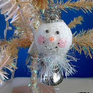 May include: A white snowman Christmas ornament with a sparkly blue and silver crown, pink cheeks, and a silver bell. The ornament is hanging from a white artificial Christmas tree.