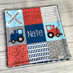May include: A blue and red patchwork baby blanket with a train theme. The blanket features two trains, one blue and one red, and the name "Nate" embroidered in white. The blanket has a variety of patterns, including railway tracks, polka dots, and stripes.