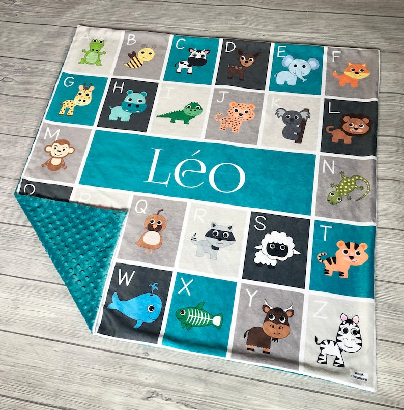 May include: A turquoise blue and gray baby blanket with a white alphabet printed on it. Each letter is paired with an animal illustration. The blanket has a soft, textured backing. The name "L&eacute;o" is printed in white on the blanket.