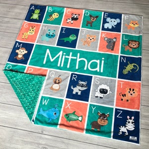 May include: A personalized baby blanket with a blue, green, gray, and orange color scheme. The blanket features an alphabet with animal illustrations and the name "Mithai" printed in white.
