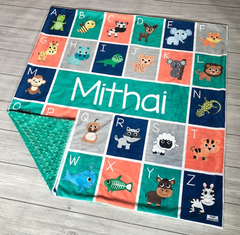 May include: A personalized baby blanket with a blue, green, gray, and orange color scheme. The blanket features an alphabet with animal illustrations and the name "Mithai" printed in white.