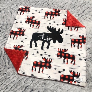 May include: A white baby blanket with a red and black buffalo plaid moose pattern. The blanket features a large black moose silhouette with the name "Lio" in script. The reverse side is a textured red fabric. The blanket is approximately 76 cm square.