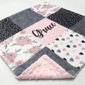 May include: A pink and gray baby blanket with a patchwork design. The blanket features a floral pattern, polka dots, hearts, and a gray fabric. The blanket is personalized with the name "Grace" in black embroidery.