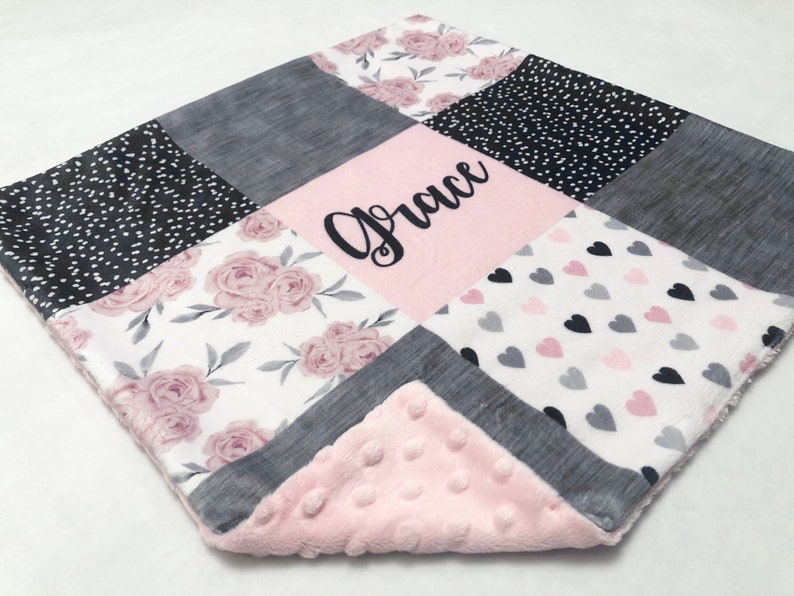 May include: A pink and gray baby blanket with a patchwork design. The blanket features a floral pattern, polka dots, hearts, and a gray fabric. The blanket is personalized with the name "Grace" in black embroidery.