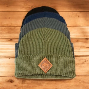 May include: A stack of four knit beanies in different shades of blue and green. The top beanie is green and features a brown leather patch with a mountain scene design.