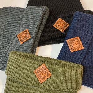 Puede incluir: Cuatro gorros de punto en diferentes colores, negro, gris, verde y azul, con un parche de cuero que presenta un dise&ntilde;o de ciervo estilizado en cada uno.