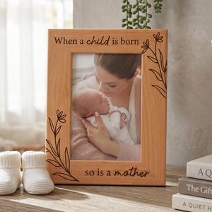 May include: A light-brown wooden photo frame with the text "When a child is born so is a mother" and floral designs. The frame contains a photo of a mother and baby. Two white baby booties are in front.