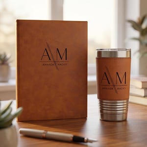 May include: A tan leather-textured notebook and a matching tan leather-wrapped stainless steel tumbler, both featuring the black printed monogram AM and the name AMANDA MACKY. A silver pen lies in the foreground.