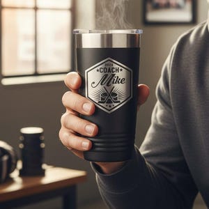 May include: A matte black stainless steel tumbler with a clear lid and steam rising from the top. The front features a white engraved hexagonal emblem with the text 2024 COACH Mike and a graphic of two crossed hockey sticks and a puck.
