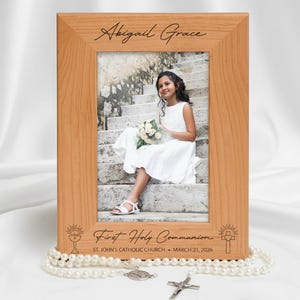 May include: A wooden photo frame with the name "Abigail Grace" engraved at the top. The frame holds a photo of a girl in a white dress. The bottom of the frame reads "First Holy Communion" and the date "March 21, 2026".
